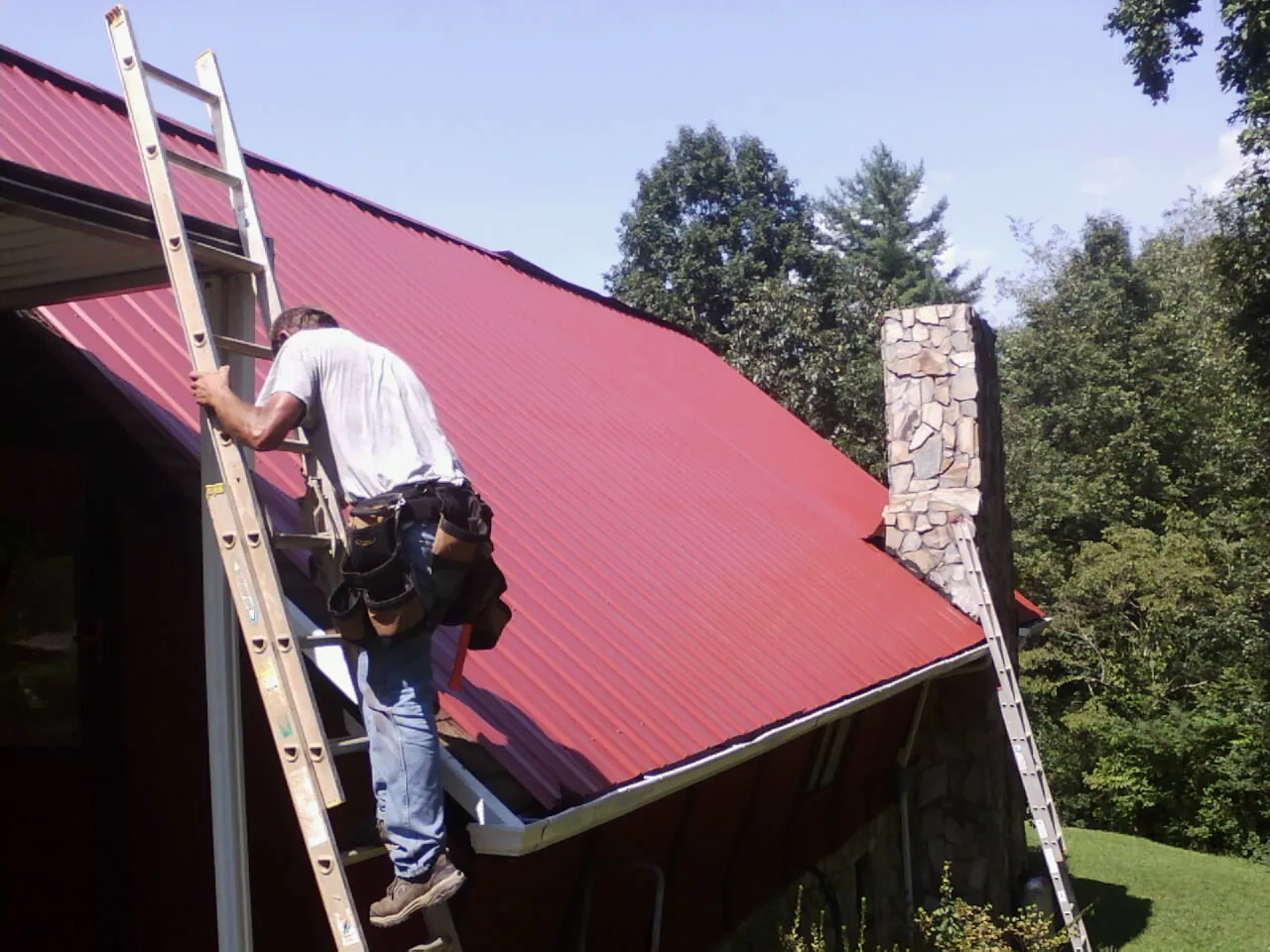 Roofer on a ladder beside a red metal roof and stone chimney — Soffit & Fascia Repair by Roofing Brothers in Susanville